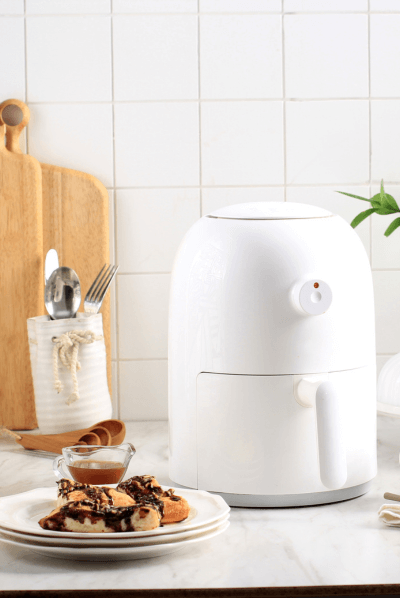 Sleek white air fryer on a kitchen counter with a plate of delicious pastries and utensils in the background.