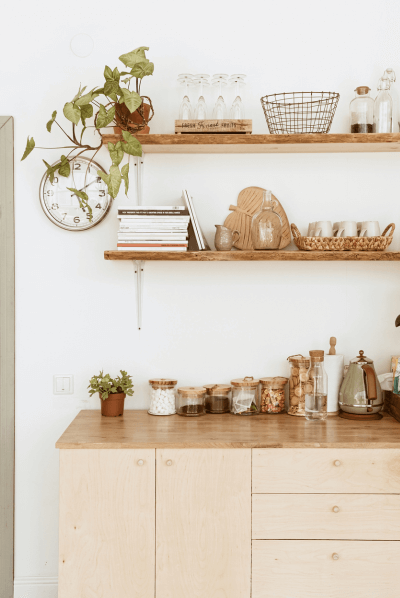 Kitchen with wooden shelves and countertop, equipped with jars, utensils, plants, and a wall clock in a clutter-free, organized manner
