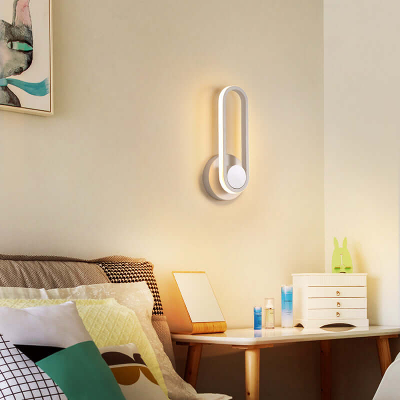 Modern oval wall lamp with warm white light installed in a cozy bedroom next to a bedside table.